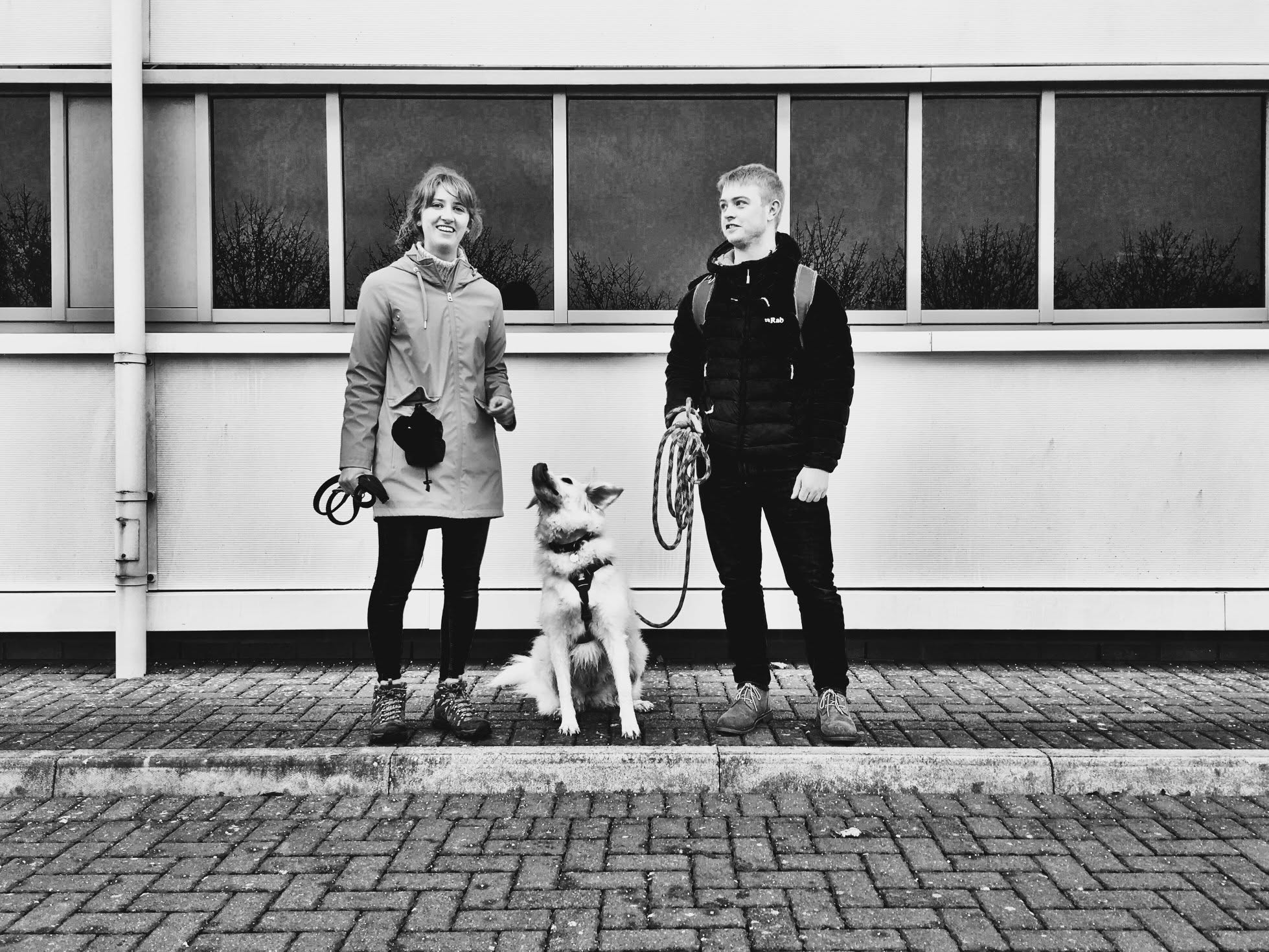 Black and white photograph of two handlers walking a white shepherd type dog on a Glasgow pavement